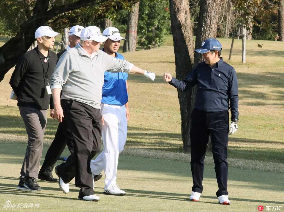 安倍晋三与特朗普站在高尔夫球场上，两人共同展示一顶白色的高尔夫球帽，帽子上绣有‘DONALD & SHINZO MAKE ALLIANCE EVEN GREATER’的字样，象征着两国同盟关系。